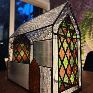 May include: A stained glass church model with a silver roof and a cross on top. The windows feature colorful diamond and triangular patterns in shades of orange, green, and red. The structure is made of glass and metal.