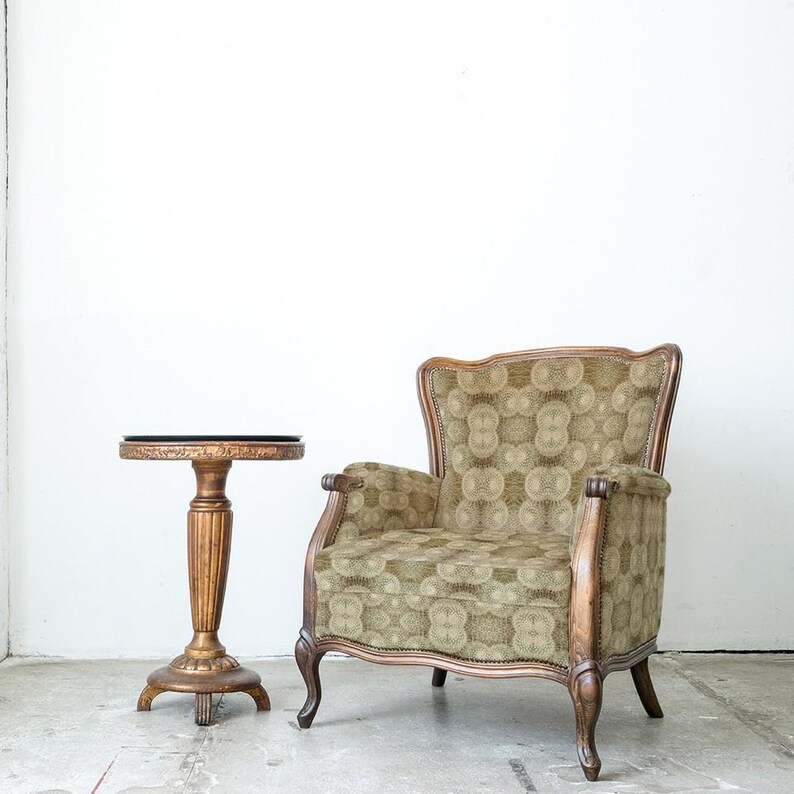 Puede incluir: Un sill&oacute;n y una mesa auxiliar antiguos. El sill&oacute;n tiene una tela verde y beige estampada con un marco de madera. La mesa auxiliar es dorada con una tapa oscura. Los muebles est&aacute;n colocados contra una pared blanca.