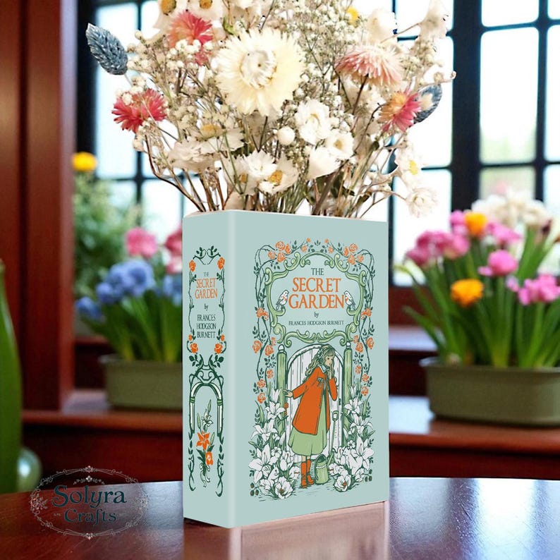 May include: A light blue rectangular vase with the cover art of "The Secret Garden" by Frances Hodgson Burnett. The vase is filled with dried flowers in shades of white, pink, and cream. The vase is sitting on a wooden surface.