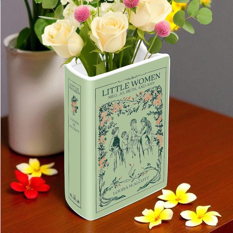 Op de afbeelding: Een lichtgroene keramische vaas in de vorm van een boek, getiteld "Little Women" met illustraties. Het bevat een boeket witte en roze bloemen. Extra bloemen liggen verspreid op het houten oppervlak.