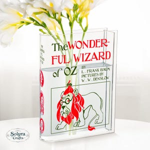 May include: A clear acrylic vase featuring the cover art of "The Wonderful Wizard of Oz" book. The vase holds white and yellow flowers. The book cover art includes a red lion illustration and the book title in red and green.