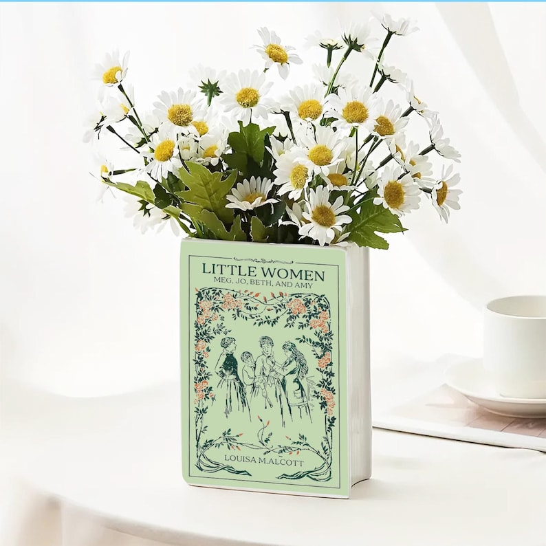 Op de afbeelding: Een witte keramische vaas in de vorm van een boek, met de titel "Little Women" en illustraties. De vaas bevat een boeket witte madeliefjes met gele centra en groene bladeren, wat een bloemstuk cre&euml;ert.