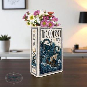 Peut inclure: Un vase en céramique rectangulaire en forme de livre intitulé The Odyssey par Homer. La face avant présente une illustration bleue d'un monstre marin attaquant un bateau. Le côté montre un trident doré. Le vase contient des fleurs blanches, roses et orange.