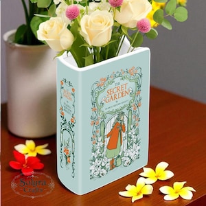 Könnte beinhalten: Hellblaues, buchförmiges Vasen-Design mit dem Titel "The Secret Garden" von Frances Hodgson Burnett. Es enthält einen Strauß cremefarbener Rosen, rosa Pomponblüten und Grünzeug. Die Vase steht auf einer braunen Holzoberfläche.