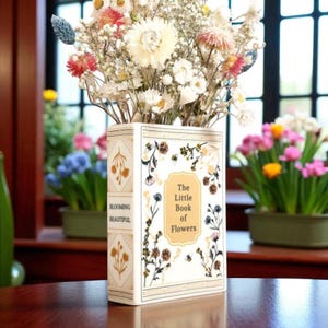 May include: A cream-coloured vase in the shape of a book, filled with dried flowers. The vase features floral and botanical illustrations and the text "The Little Book of Flowers". The spine of the book reads "Blooming Beautiful".