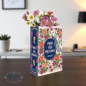 May include: A decorative vase shaped like a book, featuring the title "Pride and Prejudice" by Jane Austen. The vase is adorned with floral patterns, a peacock, and filled with colorful flowers. The vase is on a dark wooden surface.