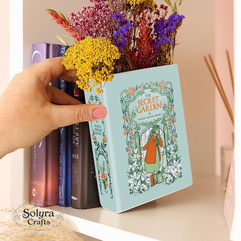 May include: A light blue ceramic vase shaped like a book, titled "The Secret Garden" with a floral illustration. The vase holds a colorful arrangement of dried flowers. The vase is on a shelf with other books.