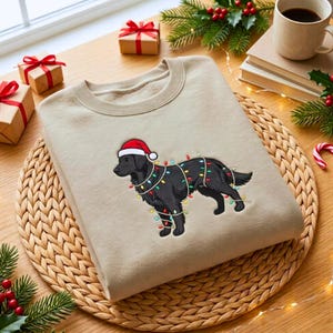 May include: A beige crewneck sweatshirt featuring an embroidered graphic of a black dog wearing a red and white Santa hat, wrapped in colorful Christmas string lights. The design is centered on the front of the garment.
