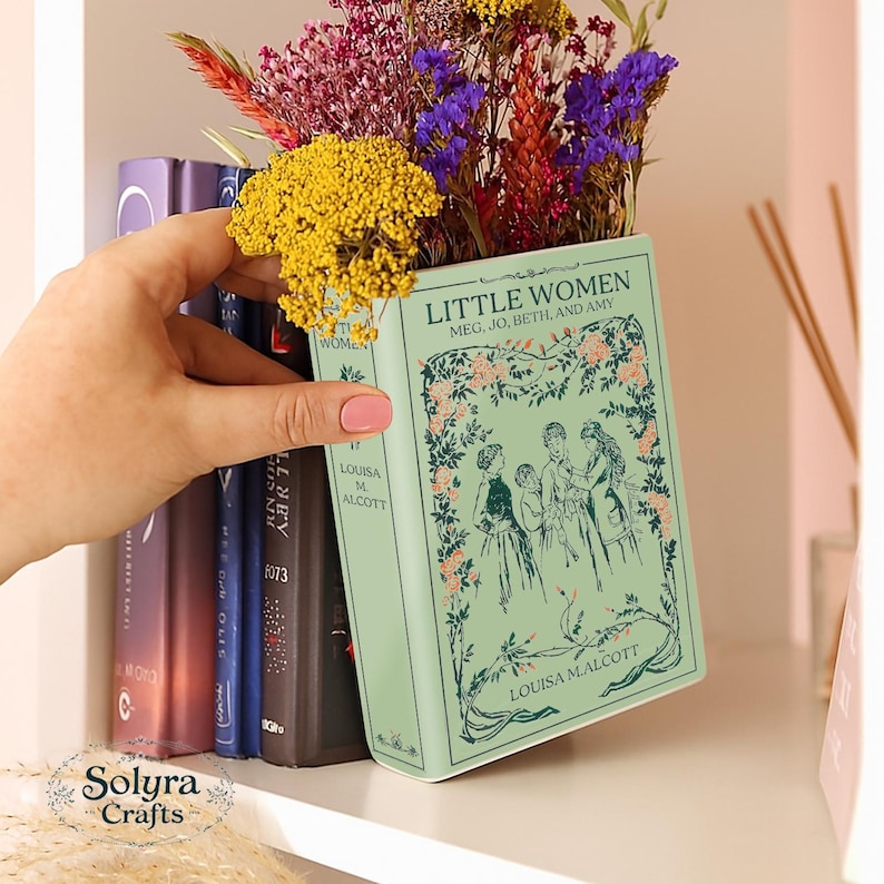 Op de afbeelding: Een lichtgroene keramische vaas in de vorm van een boek, getiteld "Little Women" van Louisa May Alcott, gevuld met kleurrijke gedroogde bloemen. De vaas staat op een plank met andere boeken.