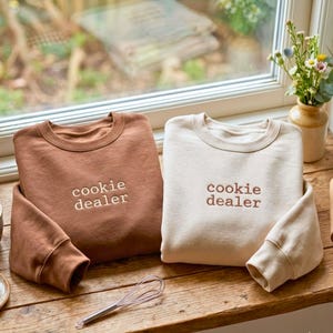 May include: Two crewneck sweatshirts, one in rust brown and one in cream, each featuring the embroidered text "cookie dealer" in a simple font. The sweatshirts are displayed side-by-side on a wooden surface near a window.