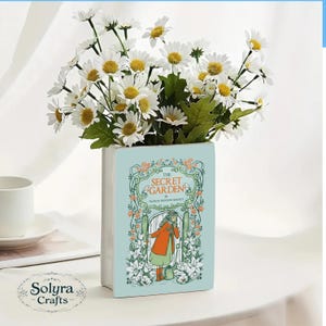 May include: A light blue ceramic vase shaped like a book, featuring the cover art of "The Secret Garden" with a girl in an orange coat. The vase holds a bouquet of white daisies with yellow centers and green leaves.