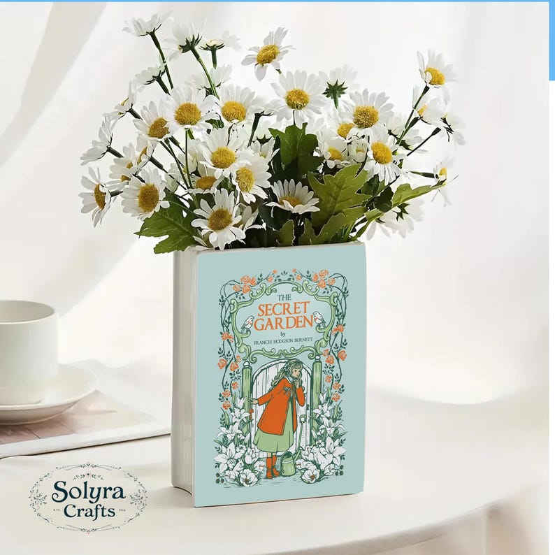 May include: A light blue ceramic vase shaped like a book, featuring the cover art of "The Secret Garden" with a girl in an orange coat. The vase holds a bouquet of white daisies with yellow centers and green leaves.