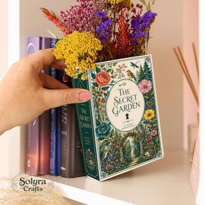 Peut inclure: Un livre évidé intitulé The Secret Garden de Frances Hodgson Burnett, orné d'une illustration florale et de jardin sur la couverture, rempli d'un arrangement vibrant de fleurs séchées jaunes, violettes et rouges.