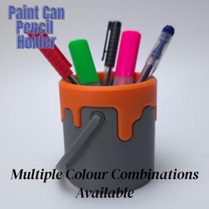 May include: A grey paint can pencil holder with an orange rim and dripping paint design. The holder contains pens and markers. Text on the holder reads "Paint Can Pencil Holder" and "Multiple Colour Combinations Available."