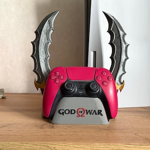 May include: A pink and black video game controller resting on a grey stand featuring the text God of War. Behind the controller are two curved, metallic-looking blades with serrated edges, positioned symmetrically to resemble the Blades of Chaos.