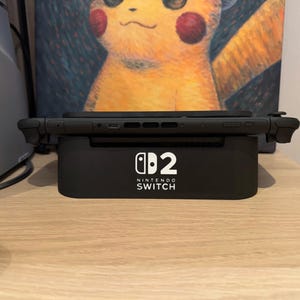 Puede incluir: Una consola Nintendo Switch negra sobre una base de carga negra. El logotipo de Nintendo Switch aparece en blanco. El fondo incluye una obra de arte de Pikachu y un dispositivo electrónico gris.