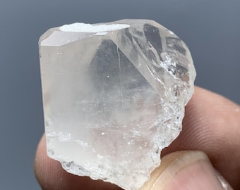 May include: A clear, faceted quartz crystal with a rough, natural edge. The crystal is mostly transparent with some internal inclusions and a white patch on top. The crystal is held in a person's hand.
