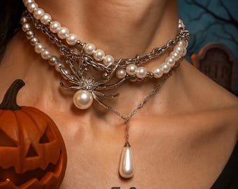 Puede incluir: Collar de varias hebras con perlas de imitación y cadenas plateadas. Un colgante de araña con una perla grande y una perla en forma de lágrima colgante. También se ve una calabaza naranja tallada y la palabra "akir".