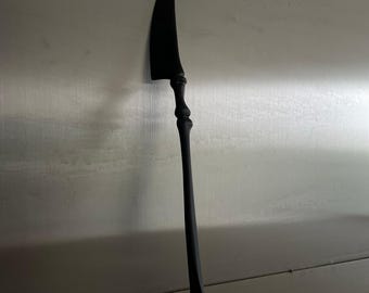 Puede incluir: Un cuchillo negro estilizado con una hoja curva y un mango largo y delgado. El mango presenta un diseño decorativo en forma de hueso. El cuchillo está sobre un fondo neutro.