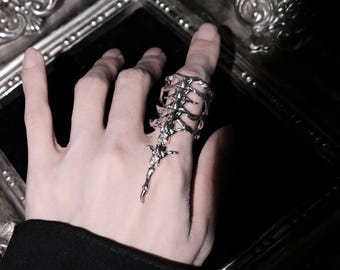 Puede incluir: Un anillo de metal plateado con un diseño alargado e intrincado que imita la estructura de una caja torácica y una columna vertebral humana. La pieza se extiende a lo largo del dedo, mostrando una superficie pulida y reflectante con líneas curvas.