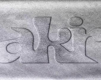 Puede incluir: Una superficie gris texturizada que presenta la palabra "aki" grabada en una fuente estilizada en minúsculas. Las letras están integradas en el material, creando un efecto de relieve con bordes suaves y redondeados y un acabado mate.
