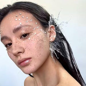Puede incluir: Retrato de primer plano de una persona con cabello y piel oscuros. La cara está adornada con adornos claros en forma de gotas y pecas. Se usa un brazalete de oreja transparente, similar al agua, en la oreja derecha, con un diseño de salpicaduras. La estética general es etérea y artística.