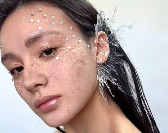 May include: A close-up portrait of a person with dark hair and skin. The face is adorned with clear, droplet-shaped embellishments and freckles. A clear, water-like ear cuff is worn on the right ear, with a splash design. The overall aesthetic is ethereal and artistic.