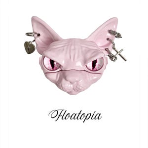 May include: A light pink Sphynx cat head figurine with intense pink eyes. The cat has silver-toned metal piercings in both ears, including a heart charm and a cross charm. The word Floatopia is printed in black script text below the cat head.