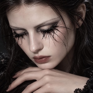 Pode incluir: Um rosto com pestanas pretas, longas e pontiagudas que se estendem para fora. Os olhos estão fechados, com sombra escura e uma cor de lábios neutra. O cabelo é escuro e ondulado, com uma peça de vestuário em renda preta texturizada.