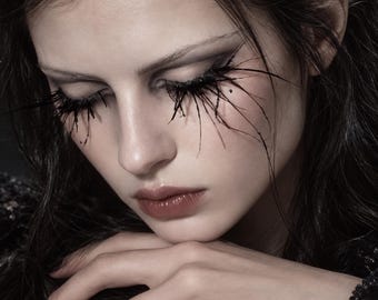 Puede incluir: Un rostro con pestañas negras, largas y puntiagudas que se extienden hacia afuera. Los ojos están cerrados, con sombra de ojos oscura y un tono de labios neutro. El cabello es oscuro y ondulado, con una prenda de encaje negro texturizada.