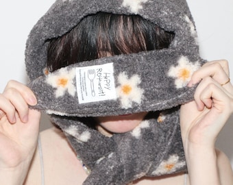 Puede incluir: Bufanda con capucha gris con diseños florales blancos y naranjas. La bufanda tiene una etiqueta que dice "Happy Restaurant!". La bufanda está hecha de un material suave y difuso.