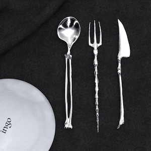 May include: Silverware set including a spoon, fork, and knife, arranged on a dark background. The spoon has a rounded bowl and a long, slender handle. The fork has three tines, and the knife has a curved blade. A white plate with the word "ingo" is in the image.
