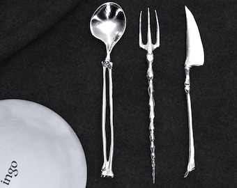 Puede incluir: Juego de cubiertos de plata que incluye una cuchara, un tenedor y un cuchillo, dispuestos sobre un fondo oscuro. La cuchara tiene un cuenco redondeado y un mango largo y delgado. El tenedor tiene tres púas y el cuchillo tiene una hoja curva. Un plato blanco con la palabra "ingo" está en la imagen.