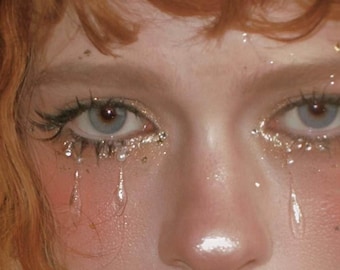 May include: Close-up of a face with blue eyes, adorned with glitter and faux tears. The skin has a rosy hue, with gold and silver glitter around the eyes. The hair is a reddish-orange color.
