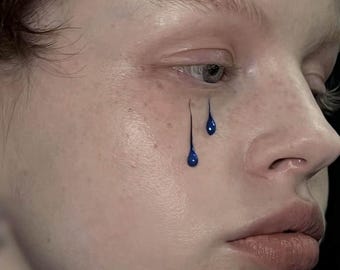 Puede incluir: Primer plano de un rostro con lágrimas de pintura azul. La piel es clara con pecas y un ligero rubor. Las lágrimas azules están estilizadas, con una línea azul oscuro que se extiende desde el ojo, terminando en forma de lágrima.