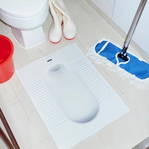 May include: A white squat toilet on a tiled floor, with a rectangular base and a curved opening. A red bucket, white boots with red soles, and a blue mop are also visible. The floor is tiled.