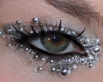 Puede incluir: Un ojo humano con iris verde y marrón, enmarcado por largas pestañas negras y decorado con numerosas esferas metálicas plateadas reflectantes de varios tamaños aplicadas alrededor de la línea de las pestañas superiores e inferiores.