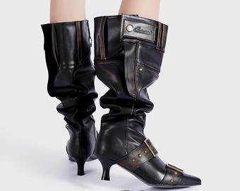 Puede incluir: Botas altas de cuero negro con punta y tacón pequeño. Las botas presentan una hebilla decorativa y detalles de correas, con un diseño ligeramente arrugado. La marca "akir" está en la parte inferior de la imagen.