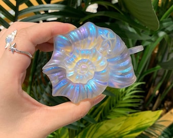 Puede incluir: Objeto decorativo iridiscente en forma de concha. El objeto tiene un diseño en espiral con un acabado nacarado, que muestra tonos de azul, morado y amarillo. Se sostiene sobre un fondo de follaje verde.