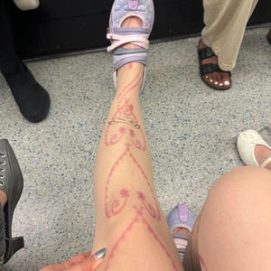 May include: A leg with a pink henna-style design. The person is wearing a lavender and pink shoe with straps. The design is on the leg and includes floral and heart-shaped patterns. The person is sitting on a grey surface.