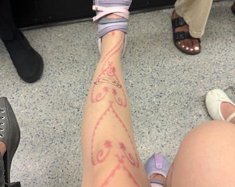 Puede incluir: Una pierna con un diseño rosa estilo henna. La persona lleva un zapato lavanda y rosa con correas. El diseño en la pierna incluye patrones florales y en forma de corazón. La persona está sentada sobre una superficie gris.