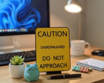 Puede incluir: Un letrero amarillo con el texto "CAUTION OVERSTIMULATED DO NOT APPROACH" está sobre un escritorio de madera. A la izquierda del letrero hay una pequeña suculenta en una maceta blanca y un objeto con forma de gato turquesa. Un bolígrafo negro y un bloc de notas también están en el escritorio.