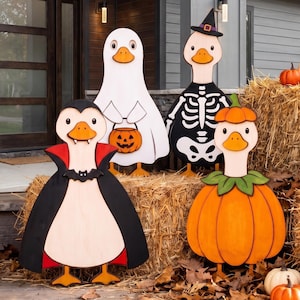 May include: Four wooden duck cutouts dressed in Halloween costumes: a vampire with a black cape, a ghost holding a jack-o'-lantern bucket, a skeleton wearing a witch hat, and a duck inside a pumpkin suit with green leaves around its neck.