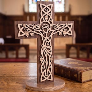 May include: A dark brown wooden cross featuring a light-colored, intricate laser-cut overlay of a figure with a crown of thorns, surrounded by Celtic knotwork patterns and grape clusters, mounted on a circular wooden base next to a book.