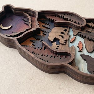 K&ouml;nnte beinhalten: Eine mehrschichtige Holzskulptur eines B&auml;ren mit einer Waldszene. Das Kunstwerk zeigt einen Halbmond, B&auml;ume, einen B&auml;ren, Fische und einen Wolf. Die Farben umfassen Braunt&ouml;ne, Blau, Orange und Lila. Die Skulptur ist ein einzigartiges Kunstwerk.