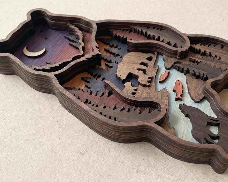 K&ouml;nnte beinhalten: Eine mehrschichtige Holzskulptur eines B&auml;ren mit einer Waldszene. Das Kunstwerk zeigt einen Halbmond, B&auml;ume, einen B&auml;ren, Fische und einen Wolf. Die Farben umfassen Braunt&ouml;ne, Blau, Orange und Lila. Die Skulptur ist ein einzigartiges Kunstwerk.