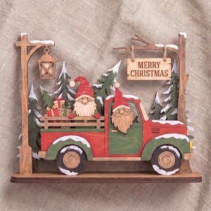 May include: A layered wooden Christmas decoration featuring a red and green pickup truck with two gnomes, one in the driver seat and one in the truck bed with gifts. A sign hanging from a tree branch reads MERRY CHRISTMAS near a street lamp.
