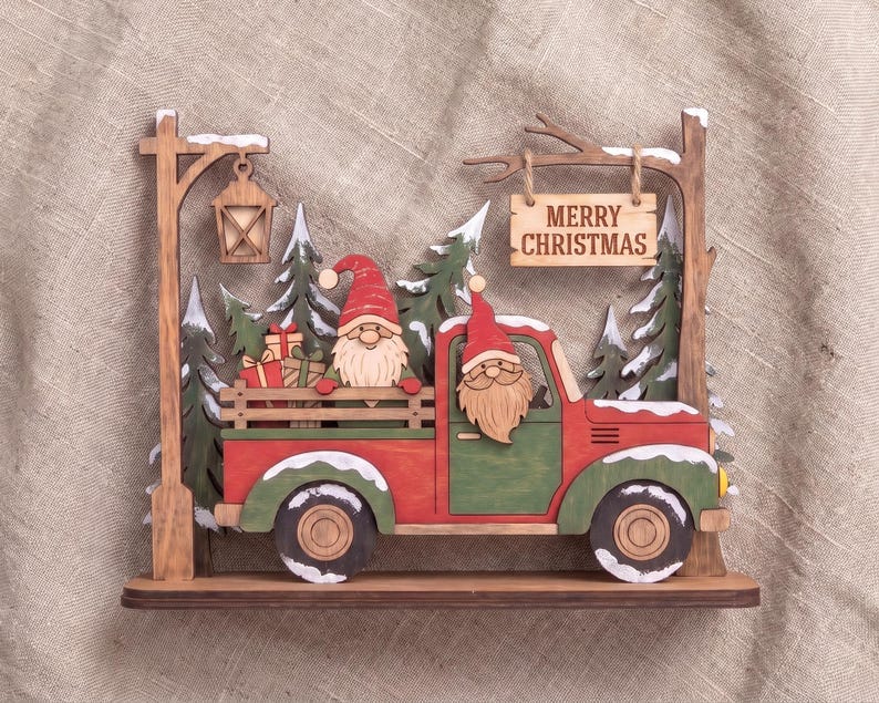 May include: A layered wooden Christmas decoration featuring a red and green pickup truck with two gnomes, one in the driver seat and one in the truck bed with gifts. A sign hanging from a tree branch reads MERRY CHRISTMAS near a street lamp.