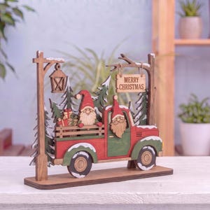 May include: A wooden Christmas decoration featuring a red and green truck carrying a gnome and gifts. The scene includes a street lamp, evergreen trees, and a hanging sign that reads Merry Christmas. The truck and trees have white snow accents.