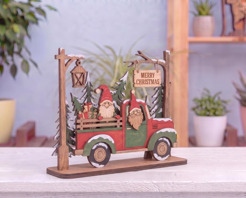May include: A wooden Christmas decoration featuring a red and green truck carrying a gnome and gifts. The scene includes a street lamp, evergreen trees, and a hanging sign that reads Merry Christmas. The truck and trees have white snow accents.